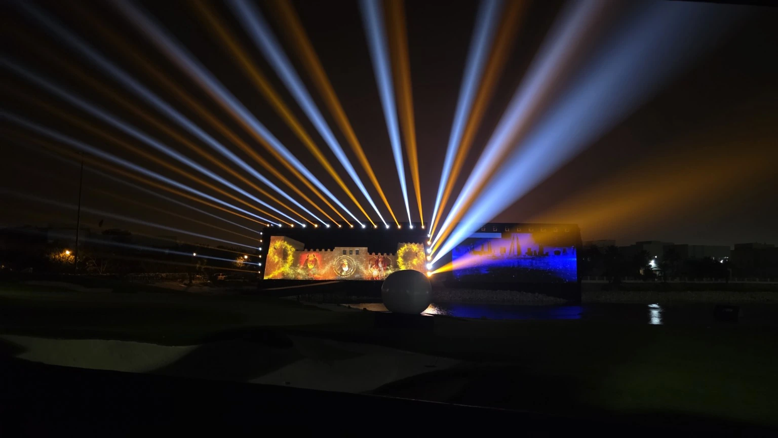Outdoor event illuminated by sweeping blue and amber light beams above vivid architectural projections